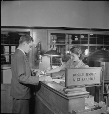 Woman behind the desk in a library stamping a book for a male patron in a suit